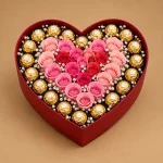 Pink and blush roses arranged in a heart with baby's breath and gold-wrapped chocolates inside a deep red gift box