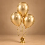Three metallic golden celebration balloons at complementary heights with matching ribbons and a decorative balloon weight